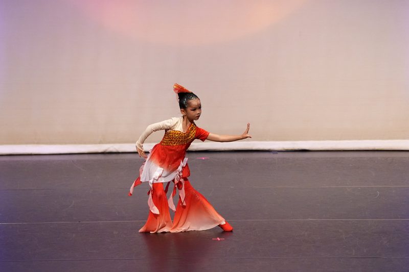 A young dancer in a red and white costume holding a poised stance on a competition stage