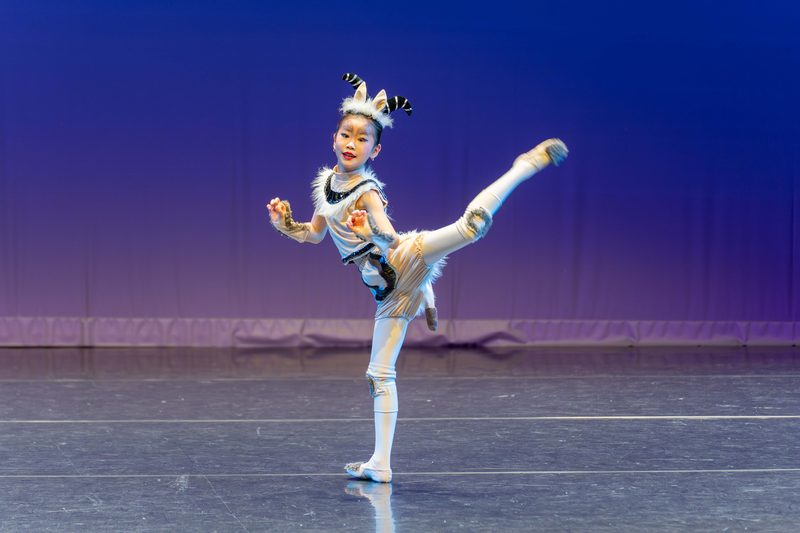 A young dancer in a horned character costume balancing on one leg in a high extension